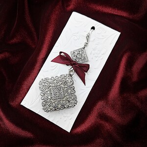 May include: Silver filigree earrings with a square design and a burgundy ribbon. The earrings are on a white card with a floral pattern.