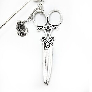 May include: Silver-toned hair clip featuring a pair of ornate scissors with a small thimble charm hanging from a chain.