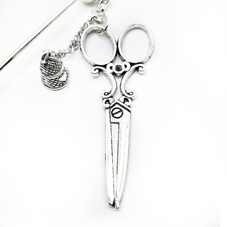 May include: Silver-toned hair clip featuring a pair of ornate scissors with a small thimble charm hanging from a chain.