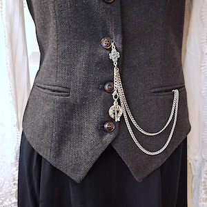 May include: A dark gray herringbone vest with brown buttons and a silver chain accessory. The chain features a four-leaf clover charm, a key charm, and a decorative chain that hangs down the front of the vest. The vest is part of a formal outfit.