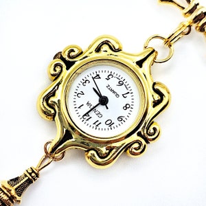 May include: A gold-tone pocket watch with a white face and black hands. The watch has a decorative, ornate frame and is attached to a gold-tone chain.