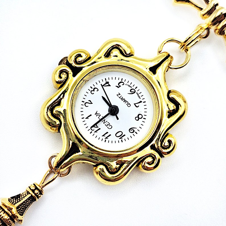 May include: A gold-tone pocket watch with a white face and black hands. The watch has a decorative, ornate frame and is attached to a gold-tone chain.