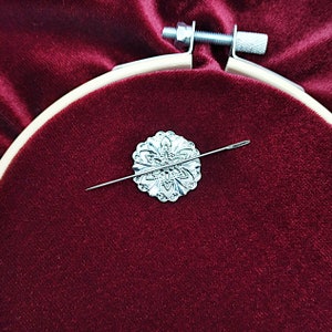May include: A silver needle threaded through a decorative silver flower-shaped needle minder, resting on a red velvet surface.