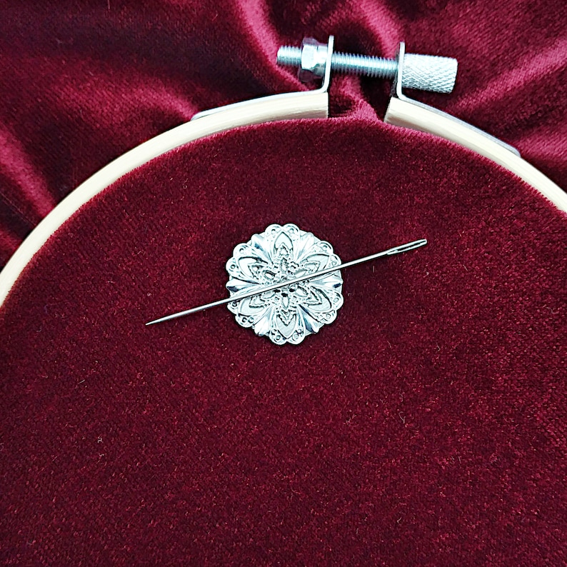 May include: A silver needle threaded through a decorative silver flower-shaped needle minder, resting on a red velvet surface.