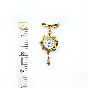 May include: Gold-toned pocket watch brooch with a white face and black Roman numeral hour markers. The watch is attached to a gold-toned chain with a decorative bow at the top. The watch is approximately 2 inches in diameter.