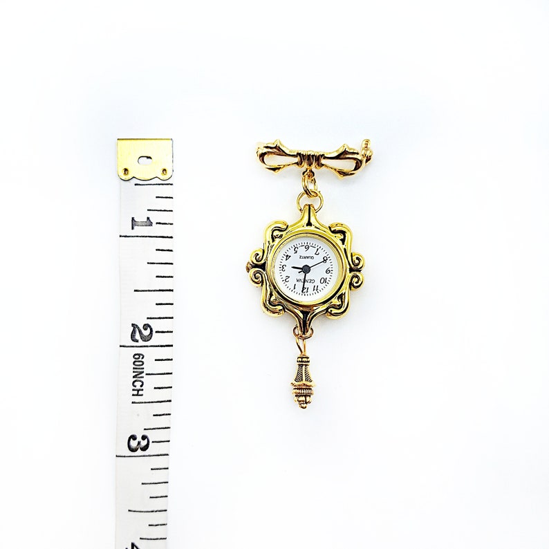 May include: Gold-toned pocket watch brooch with a white face and black Roman numeral hour markers. The watch is attached to a gold-toned chain with a decorative bow at the top. The watch is approximately 2 inches in diameter.