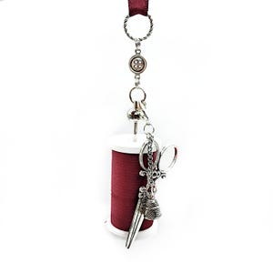 May include: A red spool of thread with a white plastic base, hanging from a silver chain with a button, scissors, and a thimble charm.