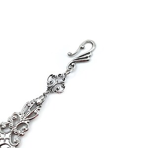 May include: Silver filigree charm with a decorative hook closure. The charm features intricate floral designs and a delicate, ornate style.