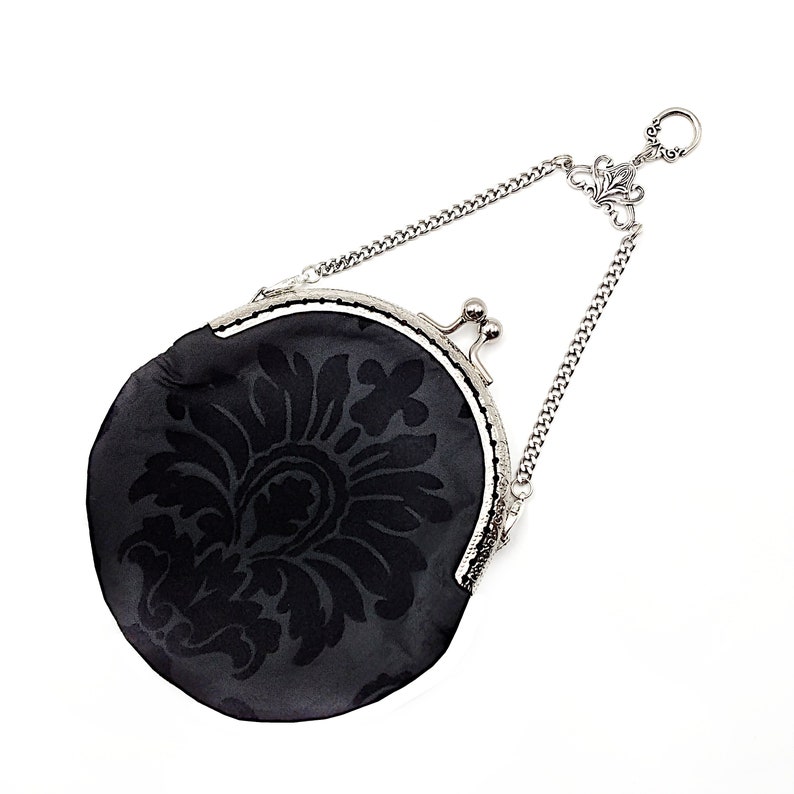 K&ouml;nnte beinhalten: Eine schwarze Stoffm&uuml;nzb&ouml;rse mit einer silbernen Kette und einem Verschluss. Die B&ouml;rse hat ein Blumenmuster.