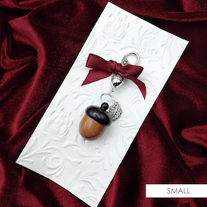 May include: A wooden acorn charm with a silver leaf and a burgundy ribbon. The charm is attached to a silver chain and is perfect for adding a touch of nature to your keychain or bag.