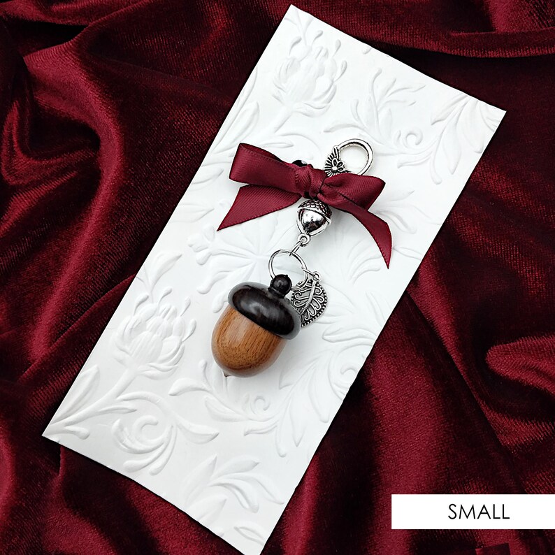 May include: A wooden acorn charm with a silver leaf and a burgundy ribbon. The charm is attached to a silver chain and is perfect for adding a touch of nature to your keychain or bag.