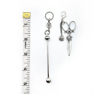 May include: Two silver charm pendants, one with a button and a ball on a chain, the other with a pair of scissors on a chain. The charms are hanging from a ruler measuring 5 inches.