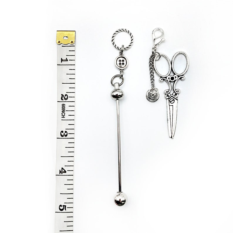 May include: Two silver charm pendants, one with a button and a ball on a chain, the other with a pair of scissors on a chain. The charms are hanging from a ruler measuring 5 inches.
