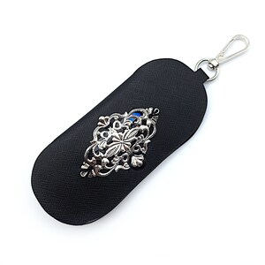 May include: A black leather key chain with a silver filigree design and a blue stone in the center. The key chain has a silver loop for attaching keys.