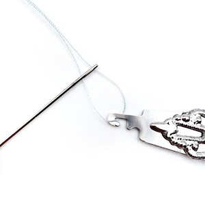 May include: A close-up of a needle threading tool. The tool is made of metal and has a hook on one end. A needle is threaded through the hook and a white thread is attached to the needle.