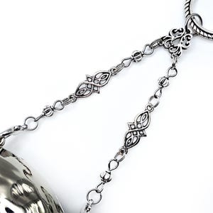 May include: Close-up of a silver-toned metal bracelet with intricate chain links and decorative filigree accents. The bracelet features a hammered metal bell-shaped charm. The design is set against a white background.