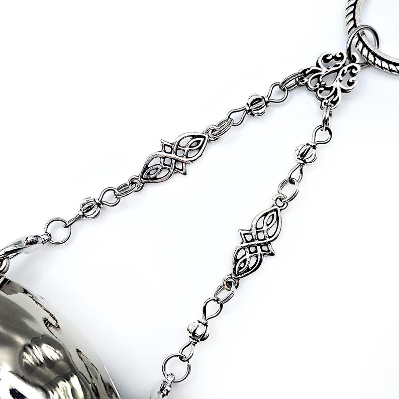 May include: Close-up of a silver-toned metal bracelet with intricate chain links and decorative filigree accents. The bracelet features a hammered metal bell-shaped charm. The design is set against a white background.