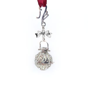 May include: A silver charm with a delicate, ornate design. The charm features a round, openwork cage with a small bell inside. A bow is attached to the top of the cage, and a loop is attached to the top of the bow.