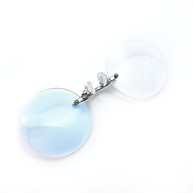 May include: A pair of round, blue-tinted reading glasses with a silver metal frame and spring hinges. The glasses are designed to clip onto existing eyeglasses.
