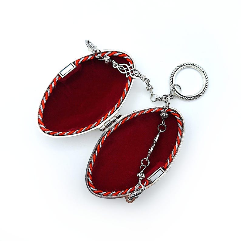 May include: Open silver-toned oval-shaped trinket box with a red velvet interior. The box is trimmed with a red and silver braided cord. A silver chain and ring are attached. The box is open, revealing the interior.