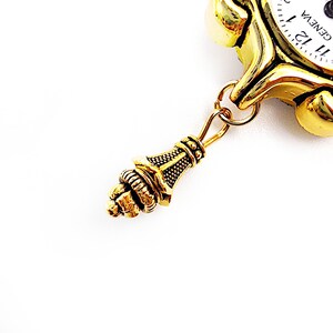 May include: Gold-toned pocket watch with a white face and black Roman numeral hour markers. The watch has a decorative, ornate, gold-toned chain with a detailed, gold-toned pendant.