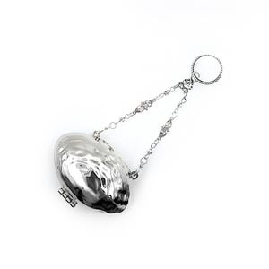 May include: A silver-colored tea infuser with a textured, oval-shaped body and a hinged clasp. It is suspended by a delicate chain with decorative links and a circular handle. The infuser is designed for steeping loose leaf tea.