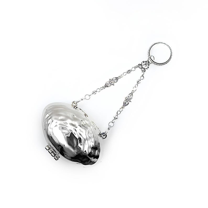 May include: A silver-colored tea infuser with a textured, oval-shaped body and a hinged clasp. It is suspended by a delicate chain with decorative links and a circular handle. The infuser is designed for steeping loose leaf tea.