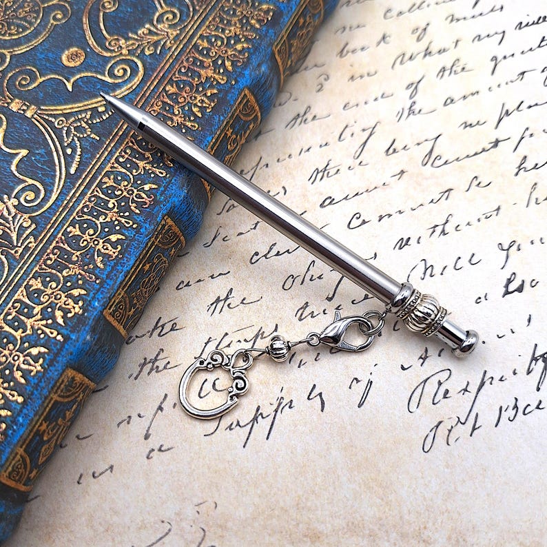 May include: A silver-toned metal knitting needle featuring a pointed tip and a decorative, ribbed end cap. Attached to the end cap is a small lobster clasp connected to a silver-toned, ornate, circular charm with intricate scrollwork details.