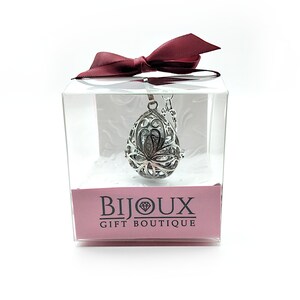 May include: A silver, teardrop-shaped aromatherapy diffuser necklace with a delicate floral design. The necklace is packaged in a clear box with a red ribbon and a pink label that reads "Bijoux Gift Boutique".