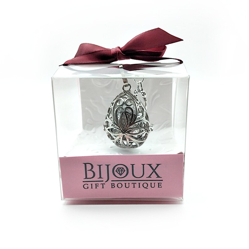 May include: A silver, teardrop-shaped aromatherapy diffuser necklace with a delicate floral design. The necklace is packaged in a clear box with a red ribbon and a pink label that reads "Bijoux Gift Boutique".