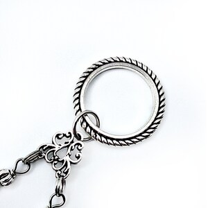 May include: Silver-toned metal jewelry piece with a large ring featuring a rope-like texture. A smaller ring connects to a decorative floral-shaped element, which is attached to a chain with small, round beads. The background is plain white.