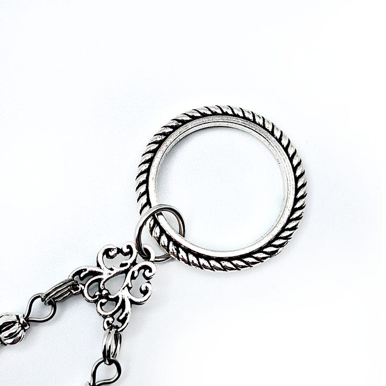 May include: Silver-toned metal jewelry piece with a large ring featuring a rope-like texture. A smaller ring connects to a decorative floral-shaped element, which is attached to a chain with small, round beads. The background is plain white.