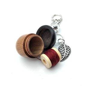 May include: A wooden acorn-shaped sewing kit with a red spool of thread, a silver leaf charm, and a silver ring with a bird charm.
