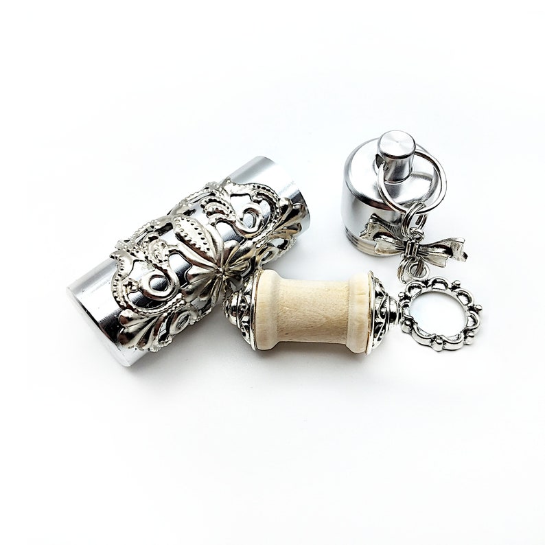 K&ouml;nnte beinhalten: Ein silberner Flaschenverschluss mit einem dekorativen Blumenmuster und einem Holzgriff. Der Verschluss hat eine silberne Kette mit einem kleinen Ring und einem Schleifenanh&auml;nger.