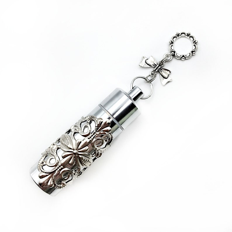 K&ouml;nnte beinhalten: Ein silberner Metallbeh&auml;lter mit einem verzierten Design, befestigt an einer silbernen Kette mit einem Schleifenanh&auml;nger und einem runden Anh&auml;nger. Der Beh&auml;lter ist etwa 5 cm lang.