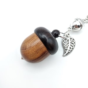 May include: A wooden acorn charm with a dark brown top and a light brown bottom. The charm is attached to a silver chain with a silver leaf pendant.