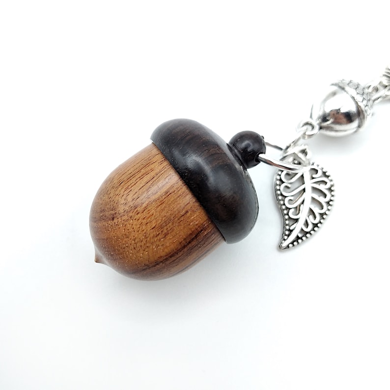 May include: A wooden acorn charm with a dark brown top and a light brown bottom. The charm is attached to a silver chain with a silver leaf pendant.