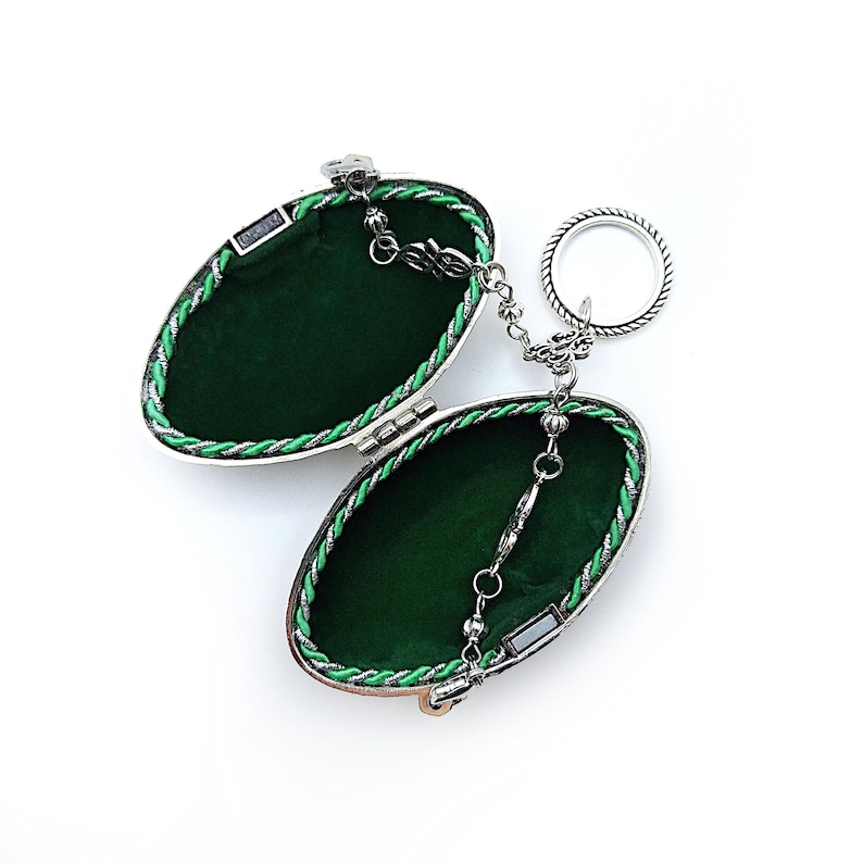 May include: An open, oval-shaped, silver-toned trinket box with a green velvet interior. The box is trimmed with green and silver rope and connected to a ring by a delicate chain. The box is on a white background.