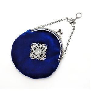 K&ouml;nnte beinhalten: Eine kleine, runde, blaue Samt-Handtasche mit einem silbernen Verschluss und einer Kette. Die Tasche hat ein silbernes Filigranmuster auf der Vorderseite.