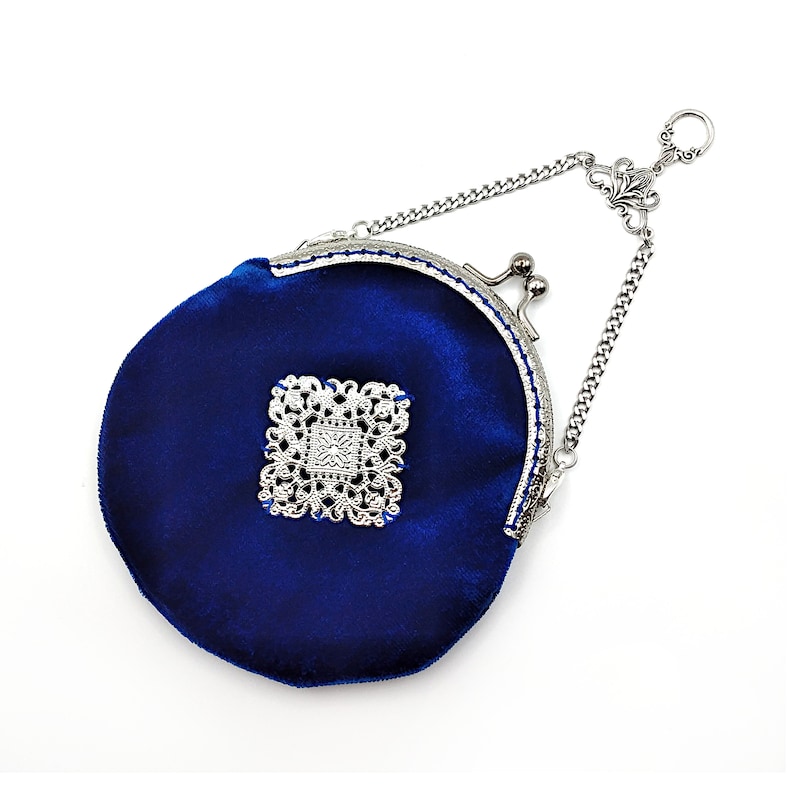 K&ouml;nnte beinhalten: Eine kleine, runde, blaue Samt-Handtasche mit einem silbernen Verschluss und einer Kette. Die Tasche hat ein silbernes Filigranmuster auf der Vorderseite.