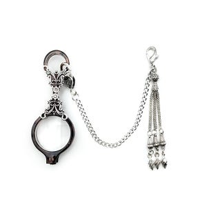 May include: A silver chain with a magnifying glass pendant and a tassel. The magnifying glass has a decorative silver frame and a black handle. The tassel is made of silver beads and has a decorative silver top.