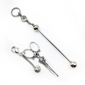 May include: Two silver-toned tie tacks. One features a button and a ball charm. The other features a pair of scissors and a ball charm.