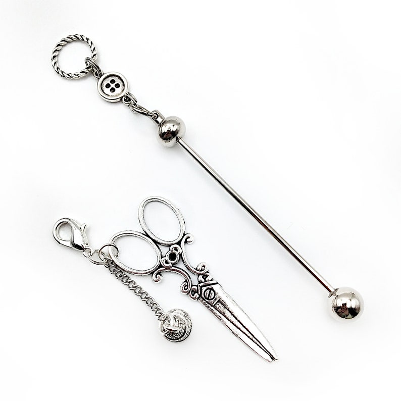 May include: Two silver-toned tie tacks. One features a button and a ball charm. The other features a pair of scissors and a ball charm.