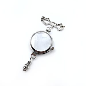 May include: A silver watch with a round face and a decorative bow-shaped pin attached to the top. The watch has a silver chain with a decorative charm at the bottom.
