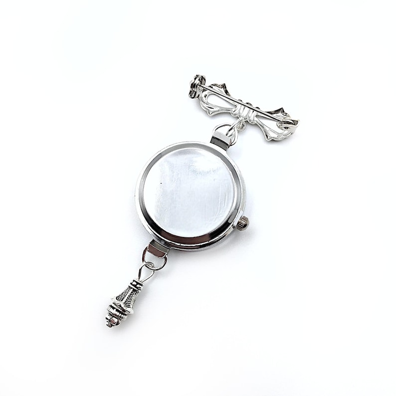 May include: A silver watch with a round face and a decorative bow-shaped pin attached to the top. The watch has a silver chain with a decorative charm at the bottom.