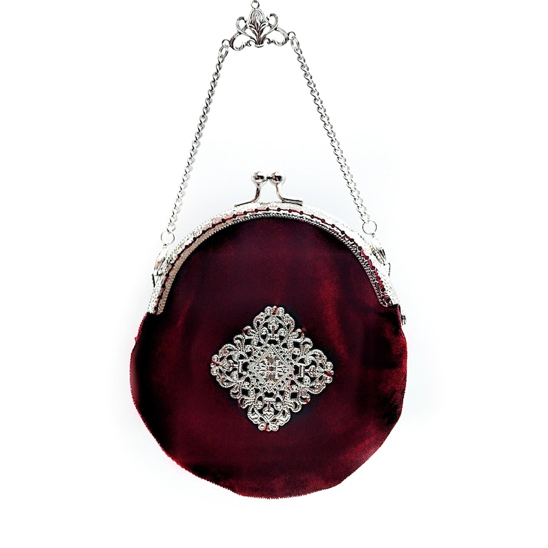 K&ouml;nnte beinhalten: Eine kleine, runde, burgunderfarbene Samt-Handtasche mit einer silbernen Kettenkette und einem silbernen Verschluss. Die Tasche hat ein gro&szlig;es, silbernes, mit Strasssteinen besetztes Design in der Mitte.