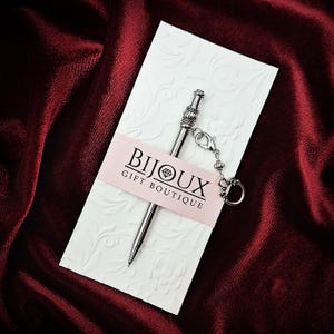 May include: A silver-toned metal pen with a decorative top and a small attached charm, secured to a white embossed card featuring the text BIJOUX GIFT BOUTIQUE printed on a light pink horizontal band across the center of the card.