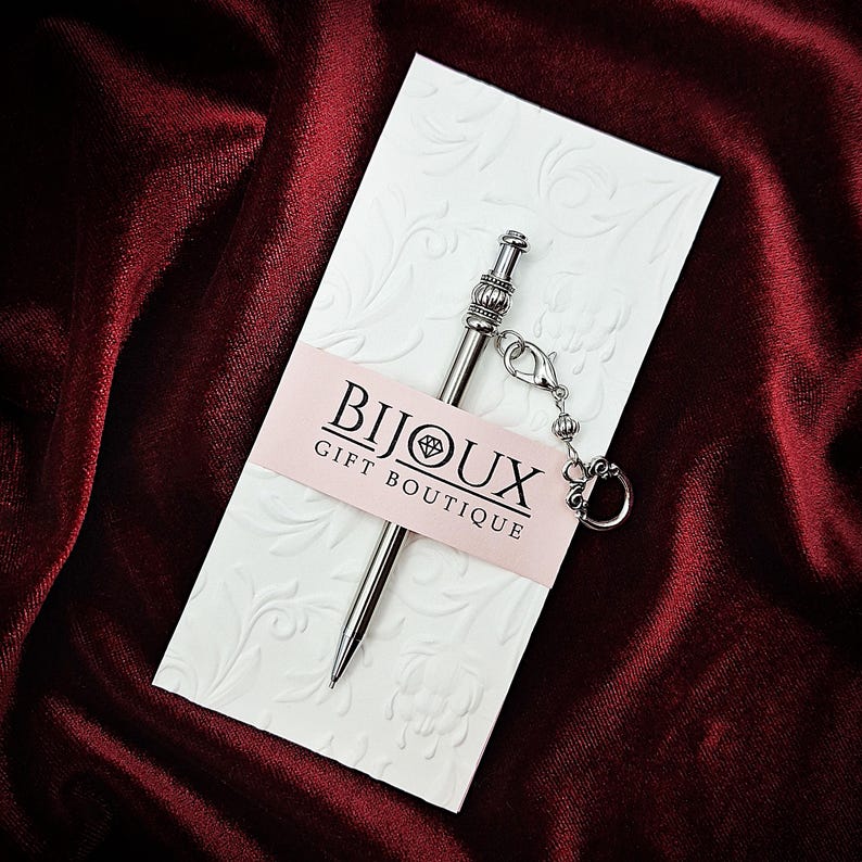 May include: A silver-toned metal pen with a decorative top and a small attached charm, secured to a white embossed card featuring the text BIJOUX GIFT BOUTIQUE printed on a light pink horizontal band across the center of the card.
