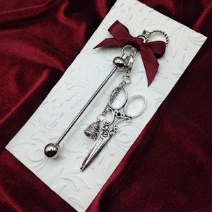 May include: A silver charm necklace with a pair of scissors, a bell, and a bead. The necklace is hanging from a white card with a red bow.