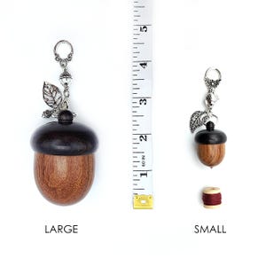May include: Two wooden acorn-shaped thread spool holders with silver acorn and leaf charms. The larger holder is 2 inches tall and the smaller holder is 1 inch tall. The words "LARGE" and "SMALL" are printed below each holder.